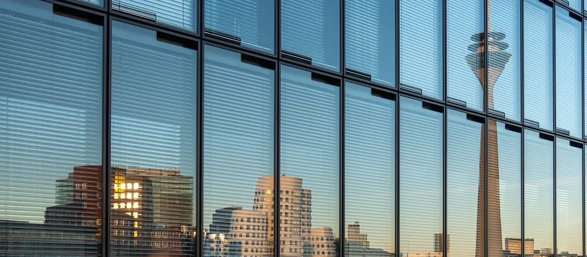 Verglaste Bürofassade mit Jalousien, in der sich der Rheinturm und die Bauten des Medienhafens spiegeln.